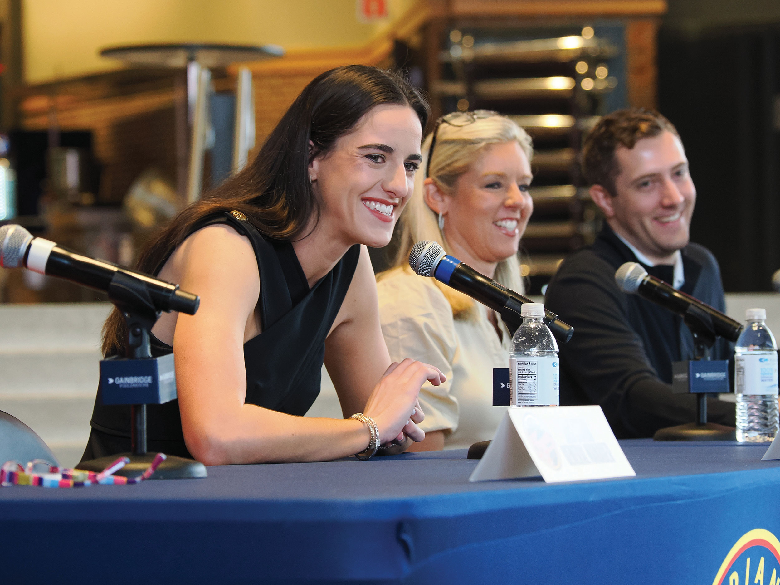 Caitlin Clark smiles as she sits beside others at a table with microphones set up in front of each person.
